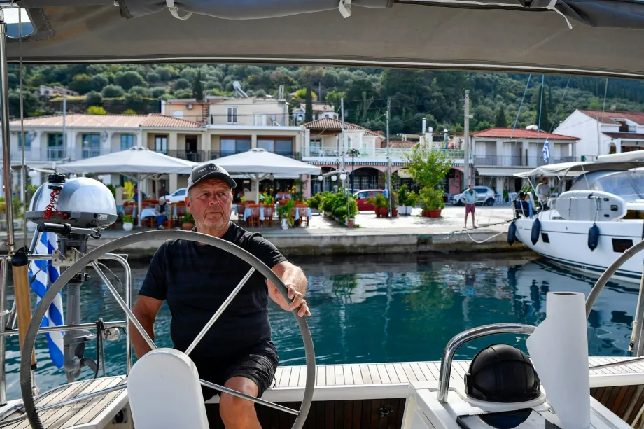 Skipper at the wheel of a sailing yacht in a harbour with waterfront buildings behind